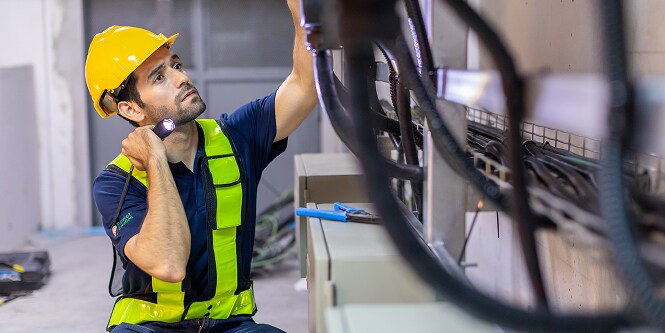 Een werknemer in veiligheidsuitrusting bekijkt elektrische aansluitingen met een zaklamp
