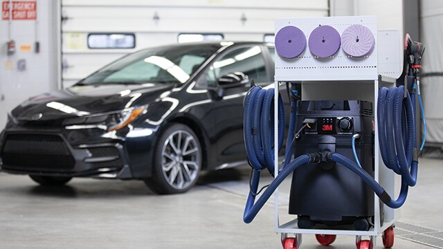 3M schuur- en stofafzuigwagen in een werkplaats, met een auto op de achtergrond
